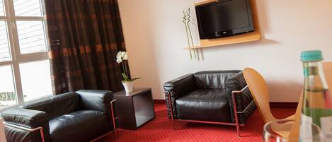 A modern living room with a red carpet, two leather sofas, a small side table with a plant, a mounted TV, and a bottle of water.