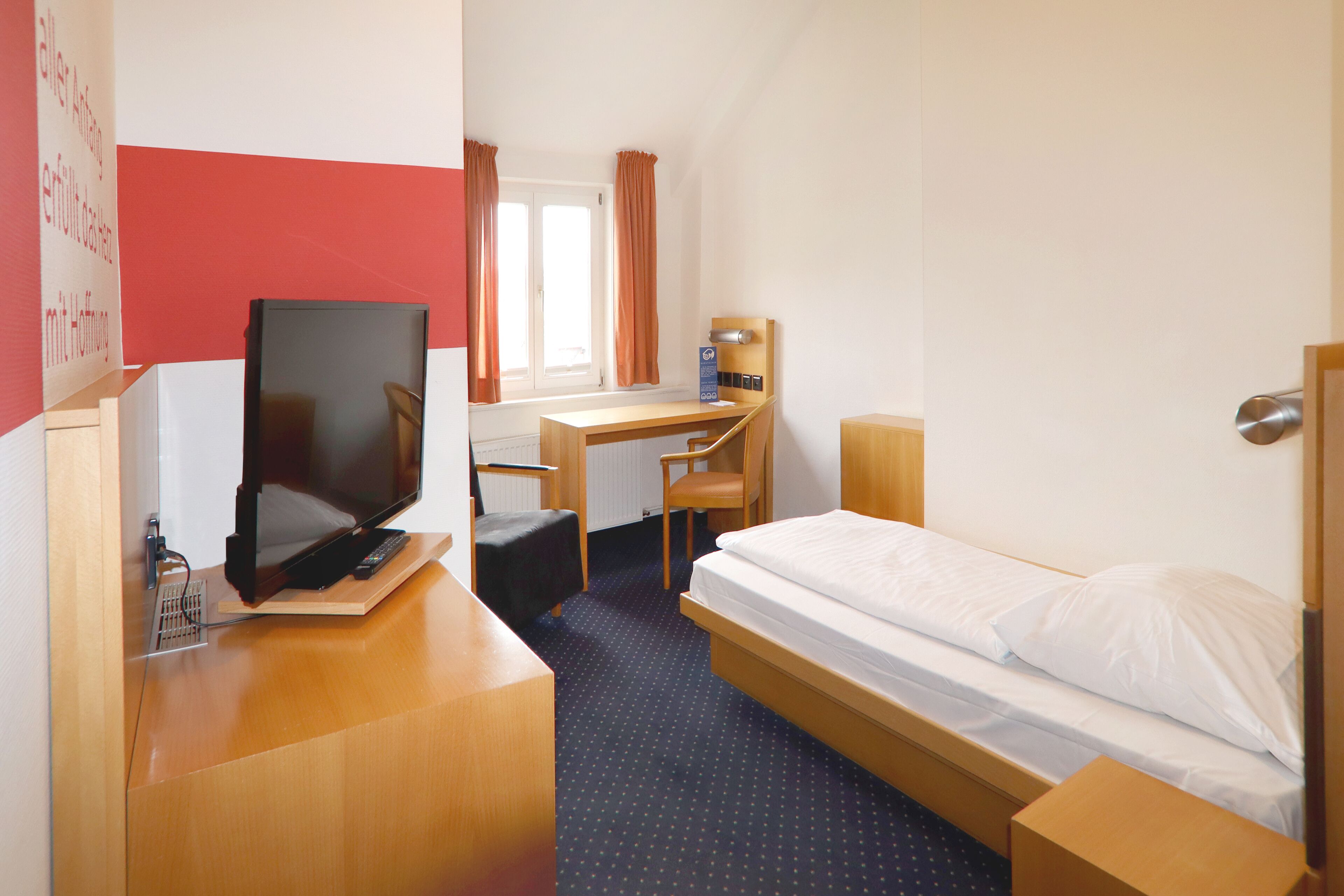 Quarto de hotel com cama, escrivaninha, cadeira e televisão.