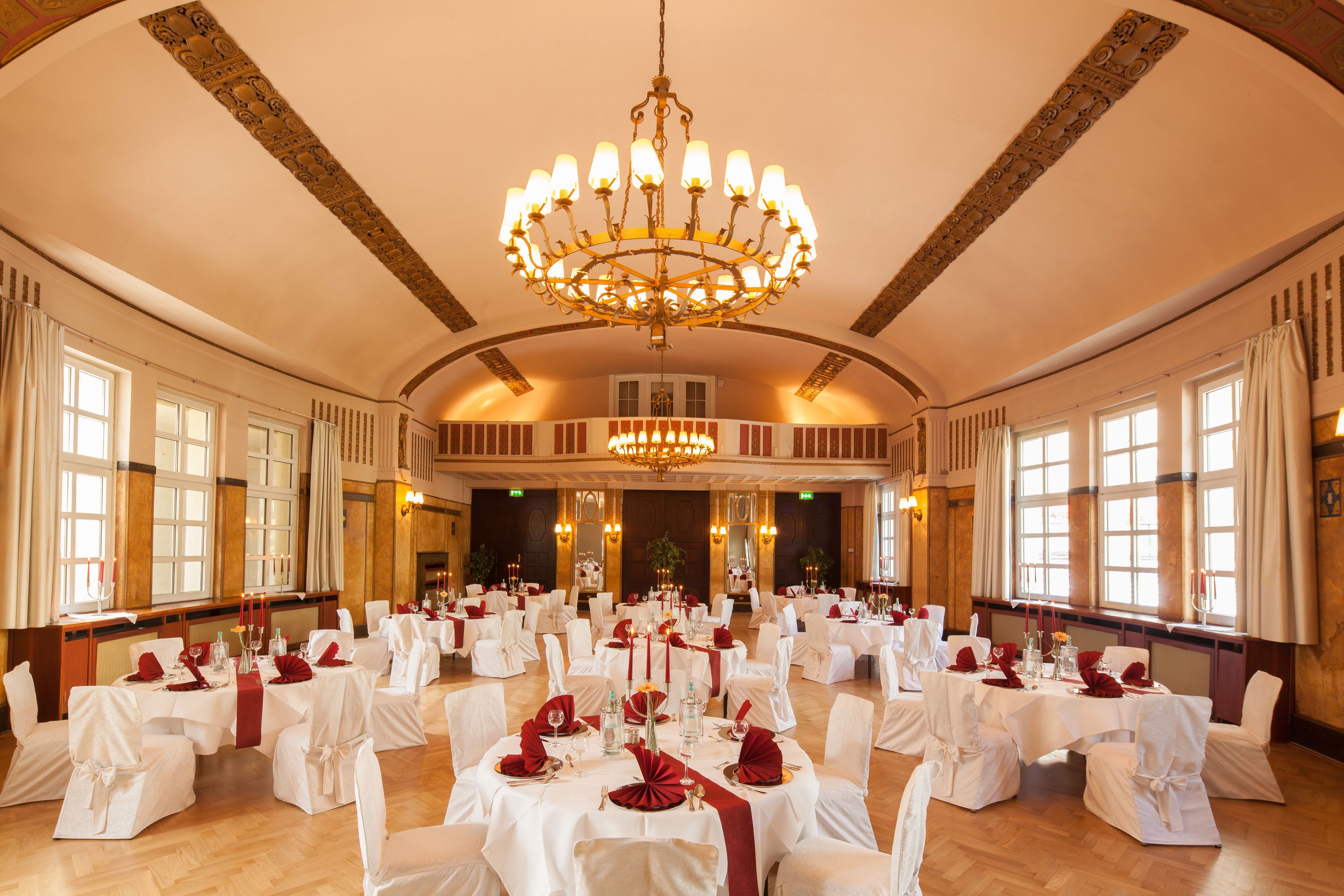 Ein Festsaal mit runden Tischen, gedeckt für eine festliche Veranstaltung, mit Kronleuchtern und großen Fenstern.