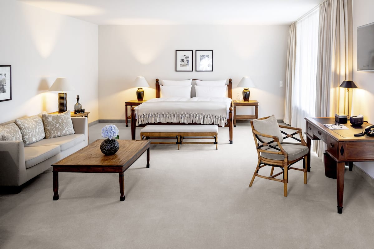 Quarto de hotel com cama, sofá, mesa de centro de madeira, cadeira, escrivaninha e duas luminárias de cabeceira.