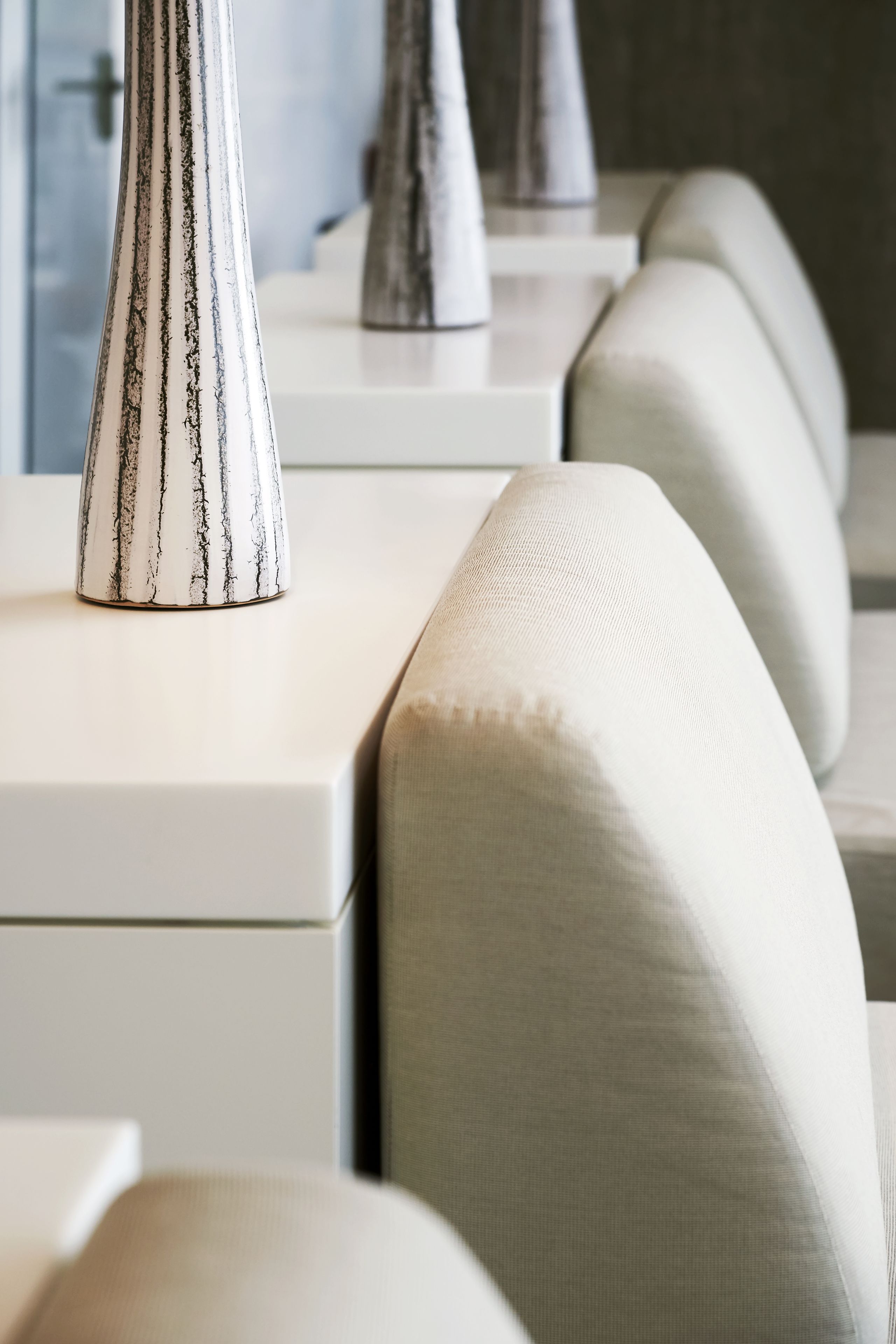 A modern hotel room with a clean, minimalist design. It features a sofa with a light fabric, a white console table, and a decorative vase.