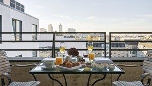 Balcón con una mesa lista para el desayuno, que incluye croissants, fruta y bebidas, con vista a un paisaje urbano.