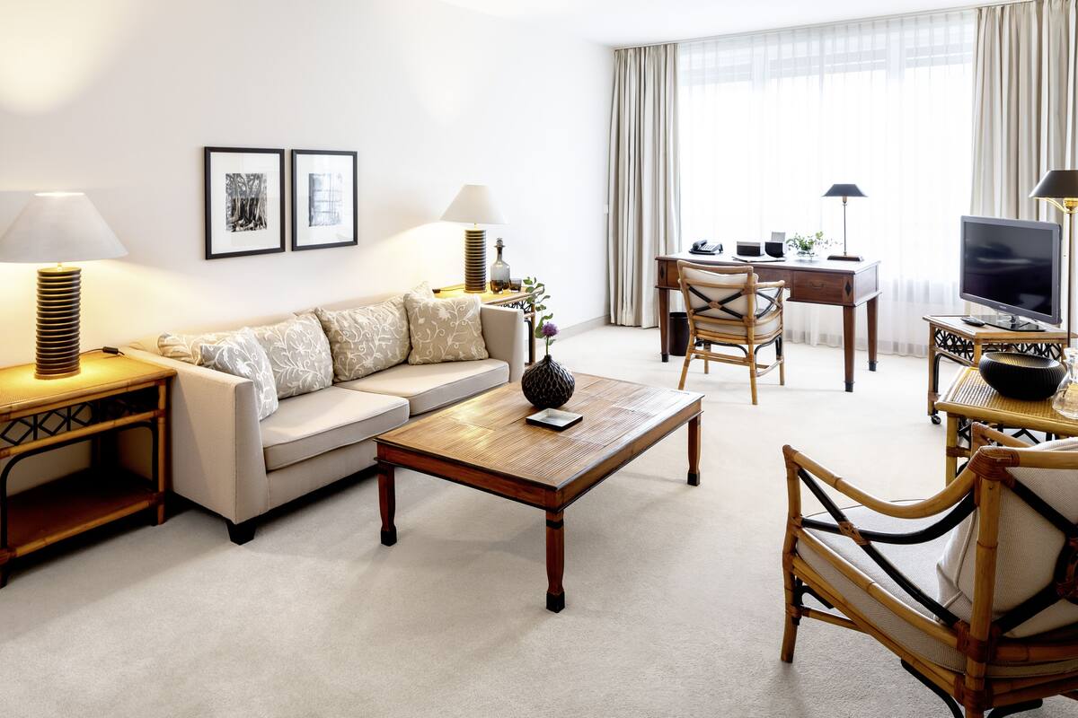 Quarto de hotel moderno com sofá, mesa de centro, mesa de jantar e cadeiras. Há duas luminárias, uma televisão e quadros nas paredes.