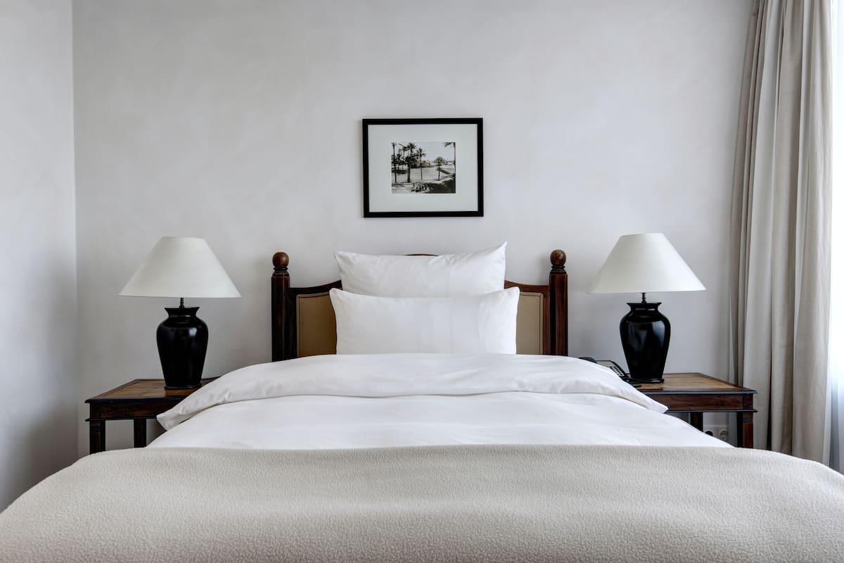 Uma cama bem arrumada com lençóis brancos, duas mesas de cabeceira com luminárias, um quadro na parede e uma janela com cortinas.