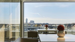 Habitación de hotel moderna con un amplio ventanal que ofrece vistas a la ciudad, una mesa con un florero y asientos cómodos.