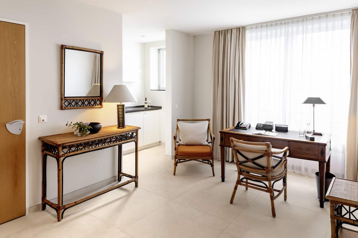 Quarto de hotel com mesa de consola de madeira, uma cadeira, secretária com computador e uma grande janela com cortinas.