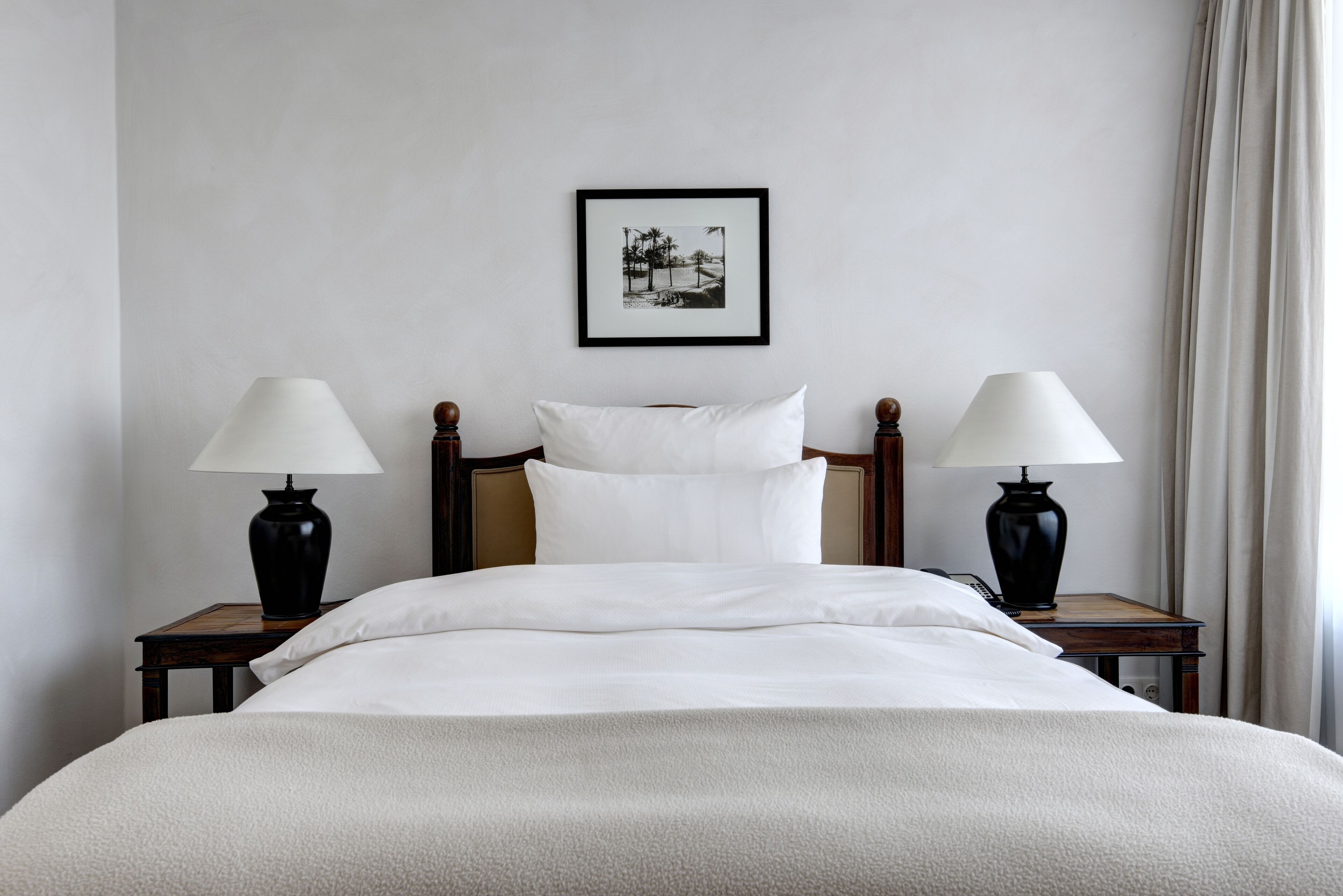 Un letto rifatto con lenzuola bianche, due comodini con lampade, un quadro appeso al muro e una finestra con tende.