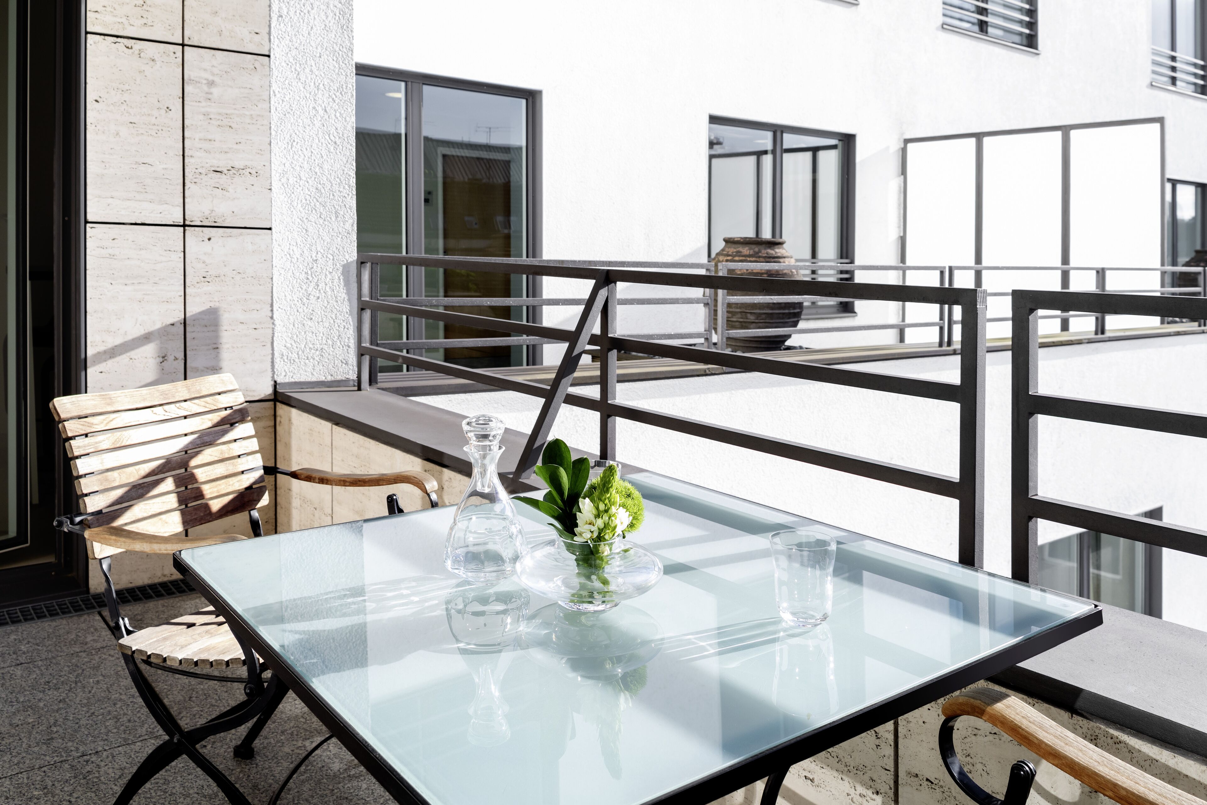 A glass-top table with a vase of flowers and a glass pitcher on a balcony with a railing.