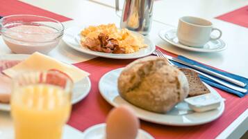 Ein Frühstückstisch mit Rührei, Speck, Toast, einem Glas Orangensaft, einem Joghurt und einer Tasse Kaffee.