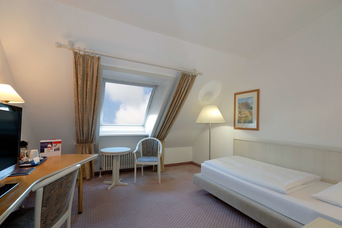 A hotel room with a bed, a desk with a computer, a chair, a lamp, a window with curtains, and a framed picture on the wall.