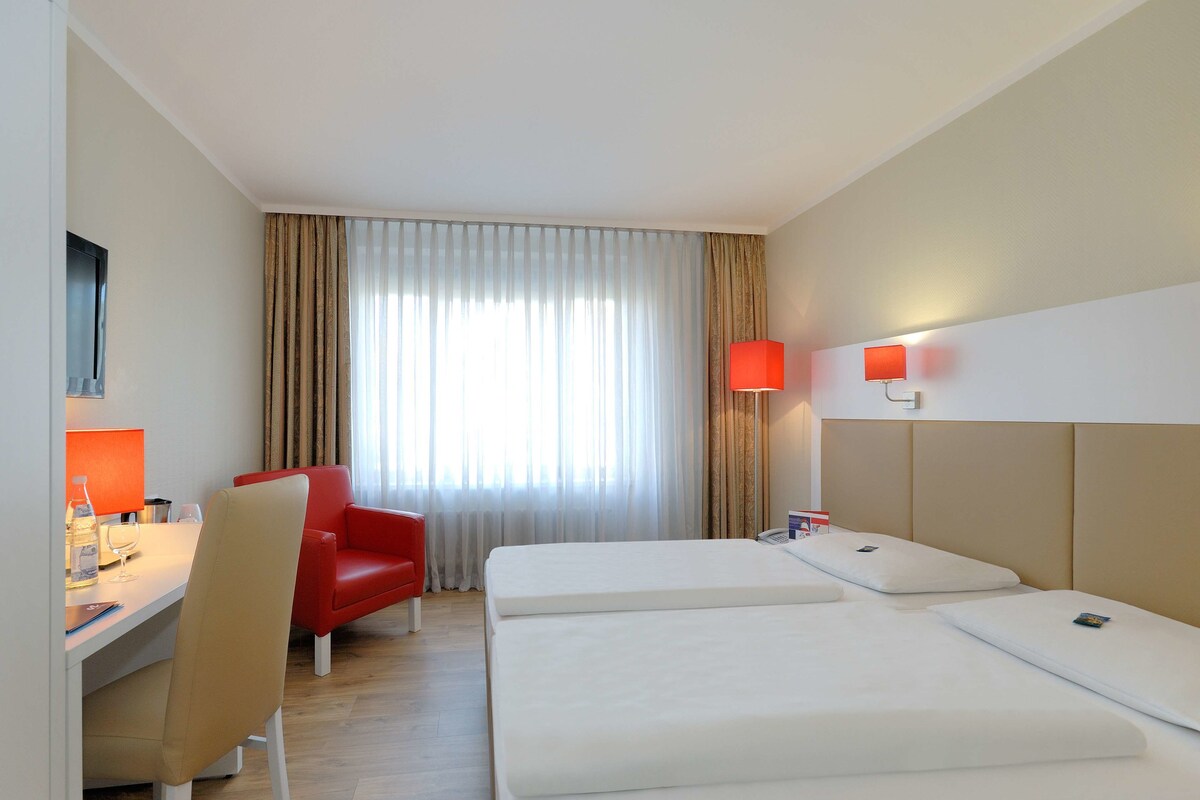 A hotel room with a bed, a desk with a chair, a red chair, a lamp, and a window with curtains.