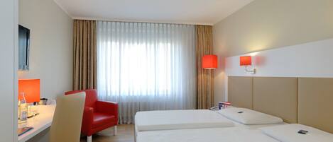 Habitación de hotel con una cama, un escritorio con una silla, una silla roja, una lámpara y una ventana con cortinas.