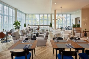 A modern restaurant with high ceilings, large windows, and a mix of seating styles including tables and chairs. - Courtyard by Marriott Dortmund (Dortmund)