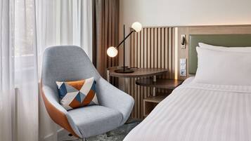 Ein Hotelzimmer mit einem Bett, einem Sessel mit gemustertem Kissen, einem Tisch mit Lampe und einem Fenster mit Vorhängen.