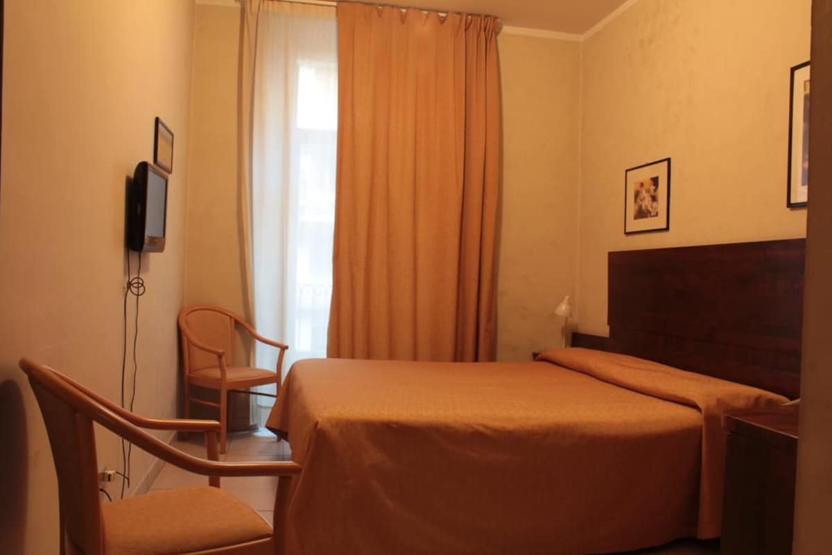 Une chambre d’hôtel avec un lit, une chaise, une télévision et une fenêtre avec des rideaux.