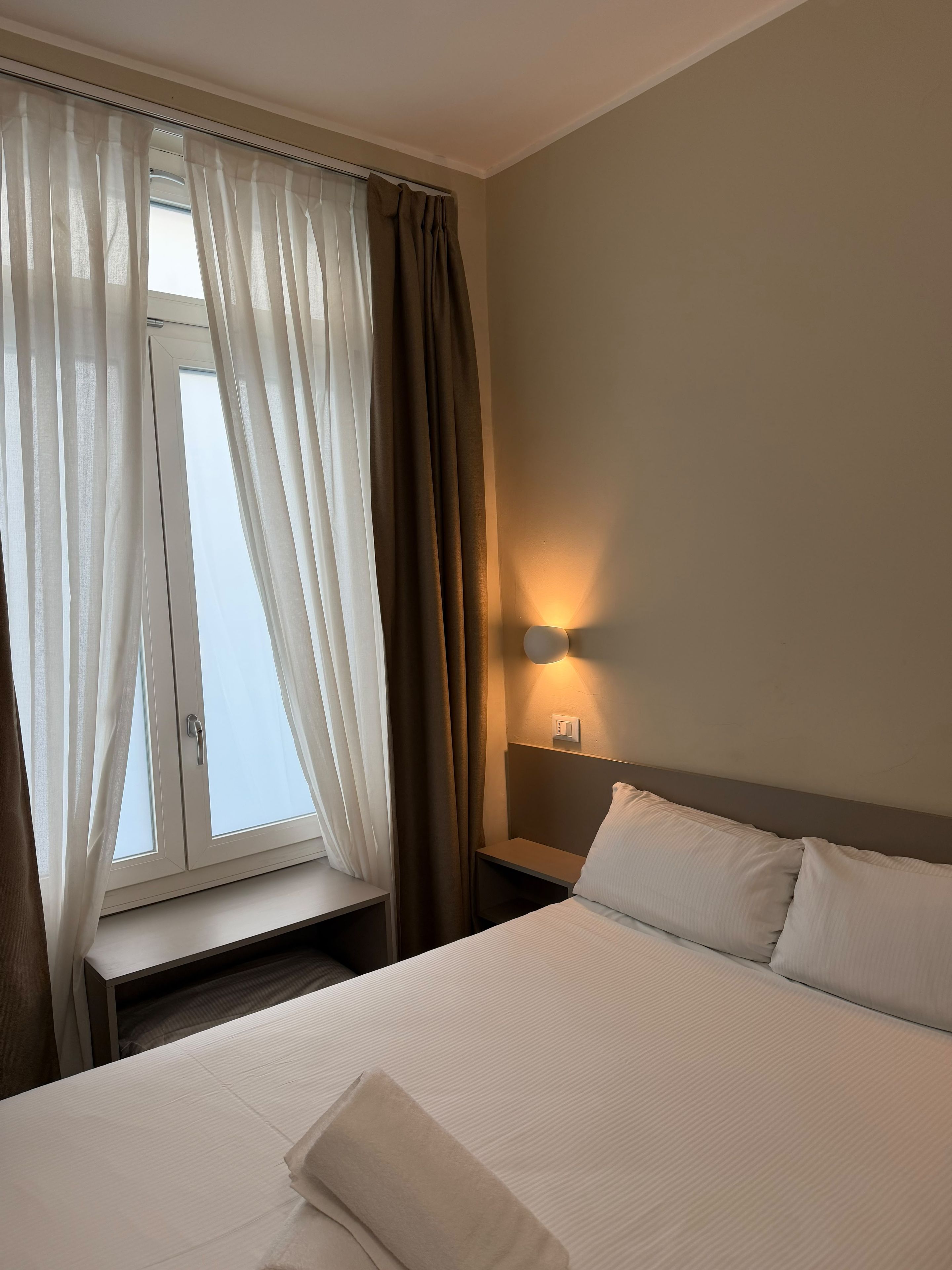A hotel room with a bed, a nightstand, a wall-mounted light, and a window with curtains.