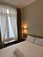 Et hotelværelse med en seng, et natbord, en lampe på væggen og et vindue med gardiner.
