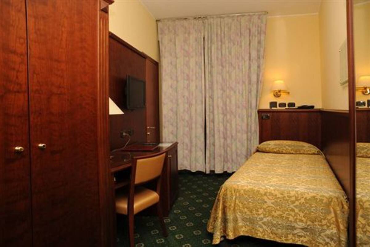 Habitación de hotel con un armario de madera, un escritorio con una silla, una cama con cubrecama estampado y una cortina.
