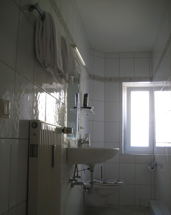 Un baño con lavamanos blanco, espejo y toallero. Una ventana permite la entrada de luz natural.