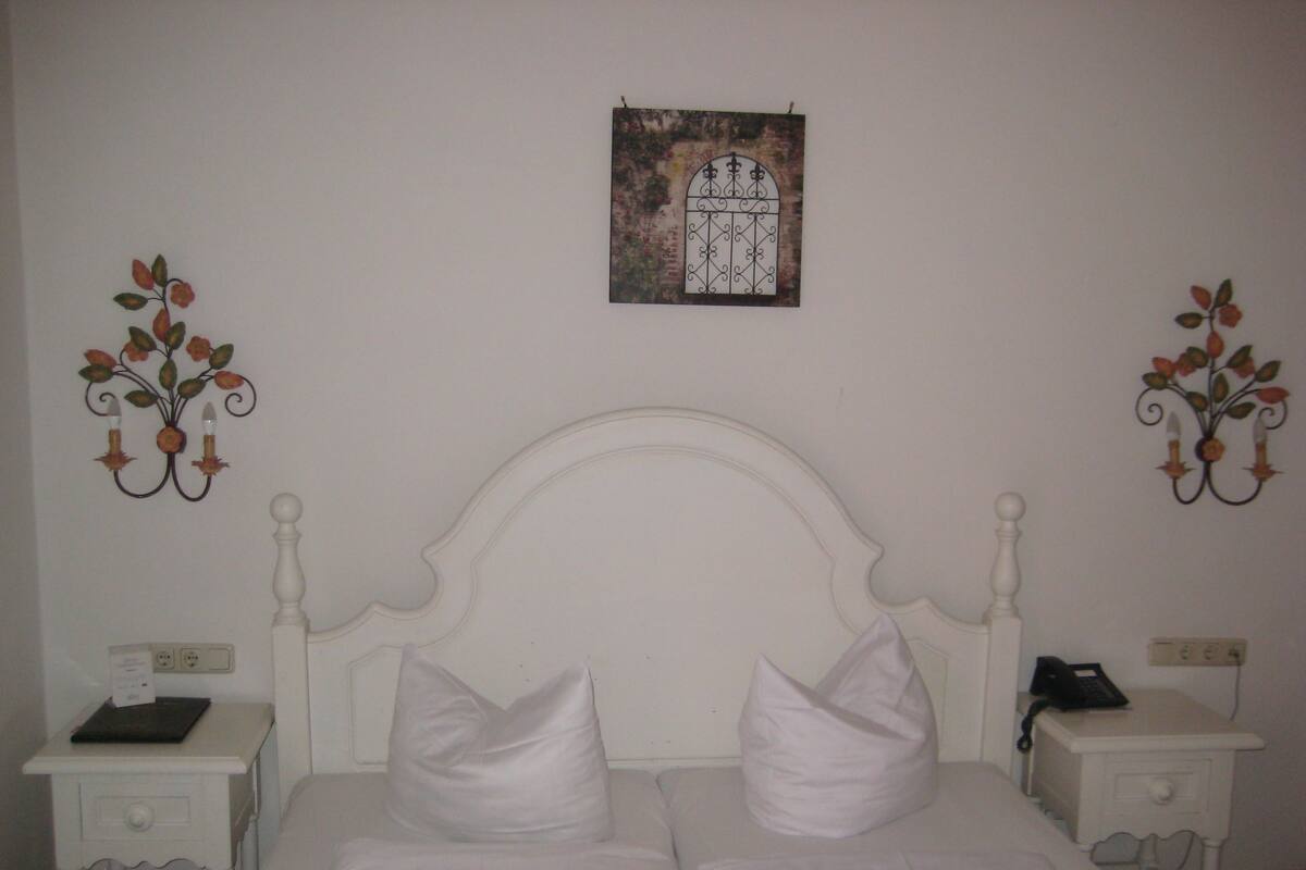 A bedroom with a white bed, two nightstands, a wall-mounted lamp, and a framed picture above the bed.