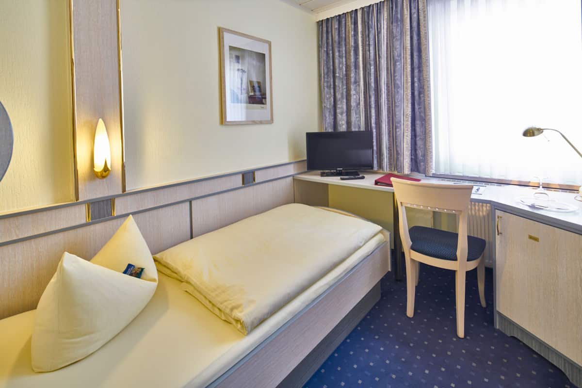 Een hotelkamer met een eenpersoonsbed, een bureau met een computer, een stoel en een raam met gordijnen.