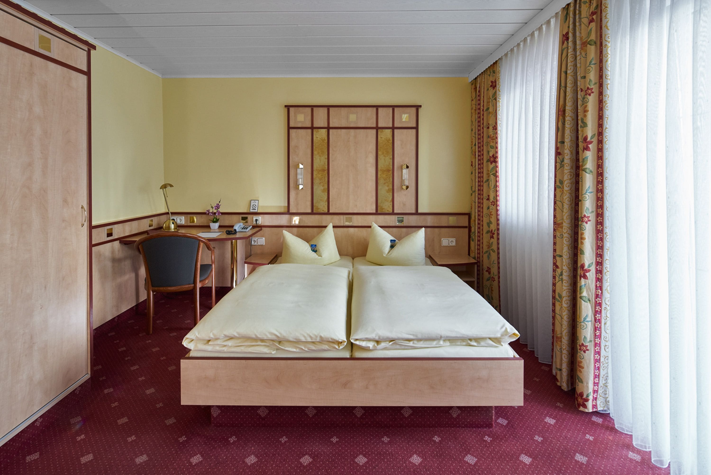 A hotel room with a large bed, a desk with a chair, a wardrobe, and curtains.