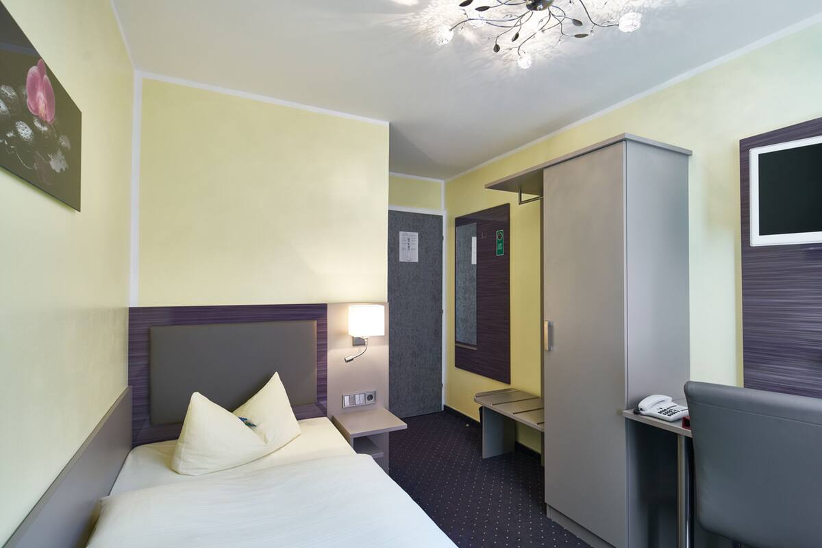 Een hotelkamer met een bed, nachtkastje, lamp, kledingkast en bureau met telefoon.