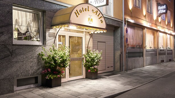 Un hotel llamado "Hotel Alfa" con una fachada clásica, una entrada acogedora y plantas en macetas a la entrada.