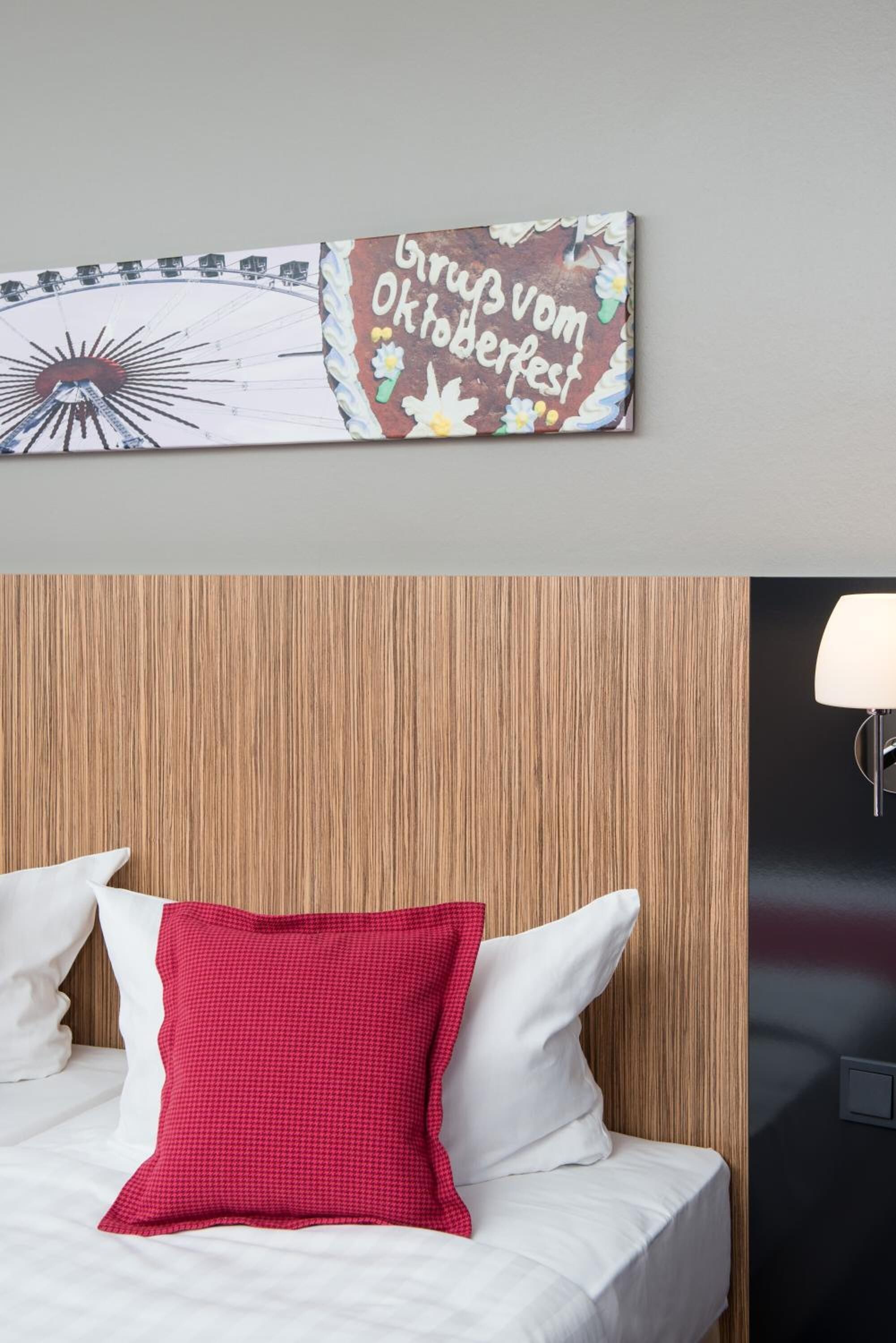 A hotel room with a wooden headboard, a bed with white and red pillows, and a wall-mounted artwork featuring a Ferris wheel and text.
