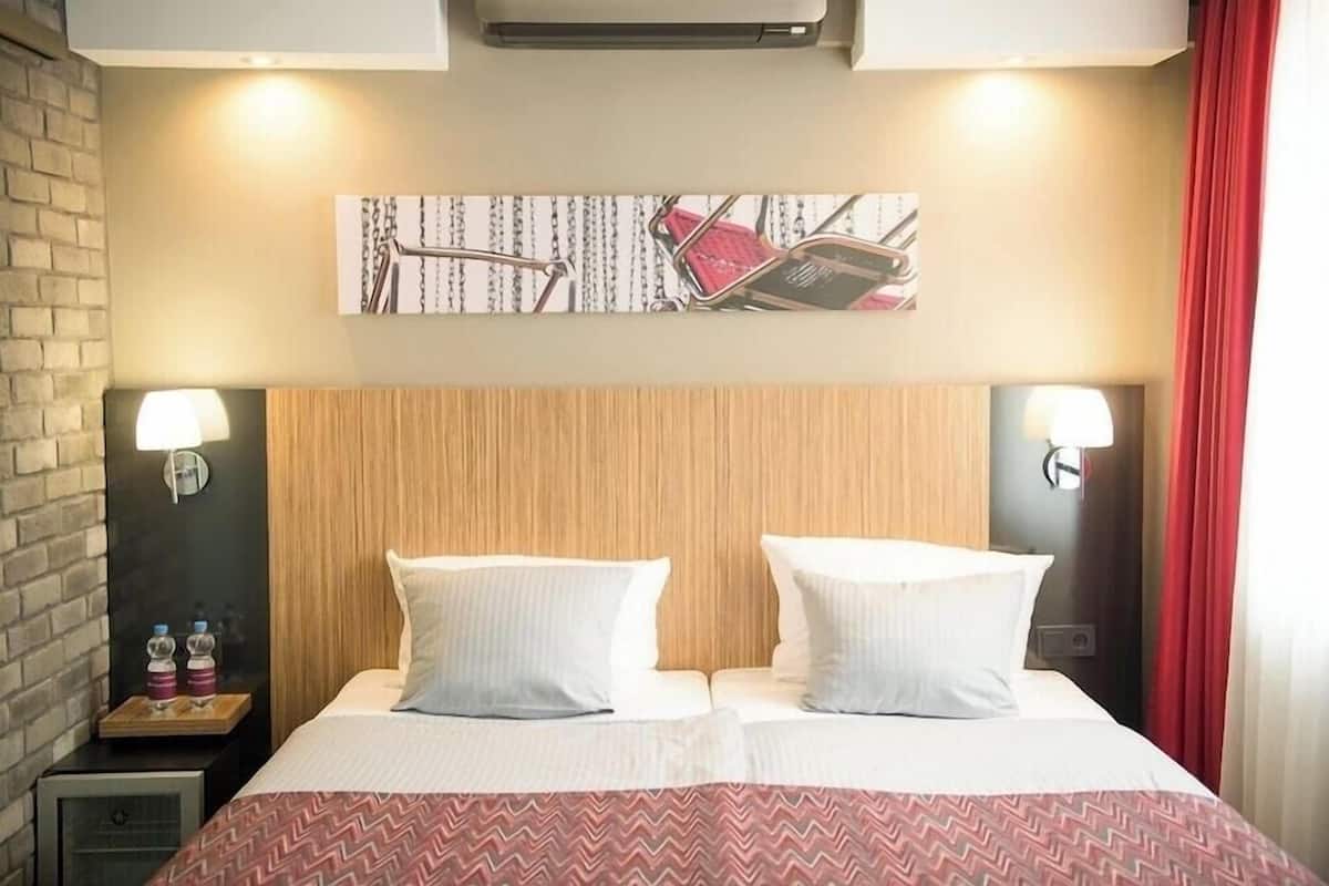 Habitación de hotel con cabecera de madera, dos lámparas de noche, cama con ropa de cama blanca, una colcha roja y blanca estampada y una obra de arte colgada en la pared.