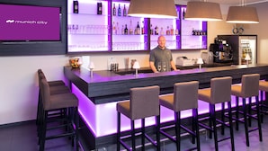A bar area with a modern design, featuring a long counter, multiple bar stools, and a variety of bottles and glasses on shelves behind the bar. - Hotel Munich City (Munich)