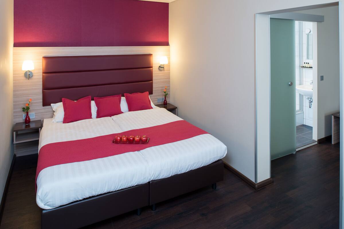 Habitación de hotel moderna con una cama grande, ropa de cama roja y blanca, una mesita de noche con una lámpara y vistas al baño.