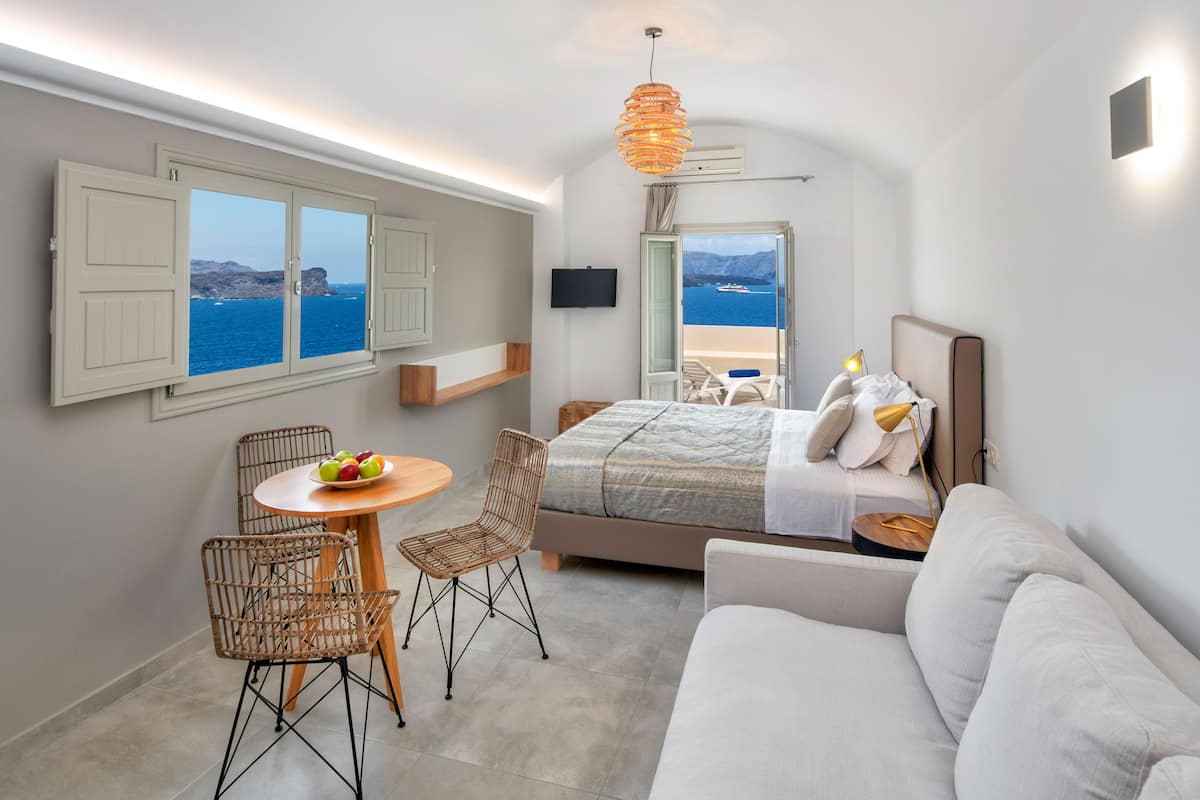 A bedroom with a bed, a sofa, a dining table with chairs, and a view of the sea.