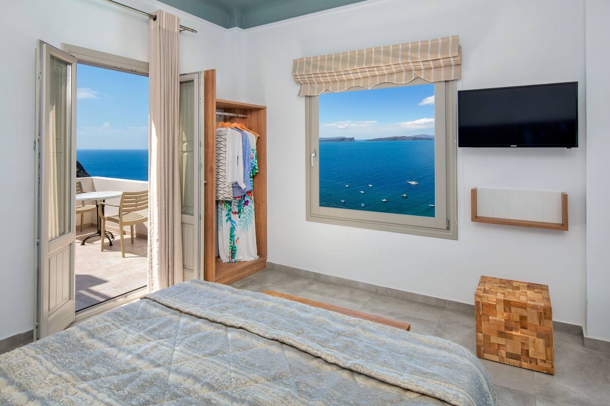 A bedroom with a bed, a TV, a window with a view of the sea, and a wooden side table.