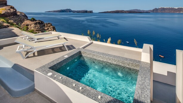 A modern outdoor pool with clear blue water, surrounded by white lounge chairs and a view of the sea and cliffs.