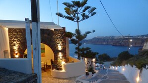 A coastal building with a stone facade, a staircase leading to a terrace, and a view of the sea and cliffs.