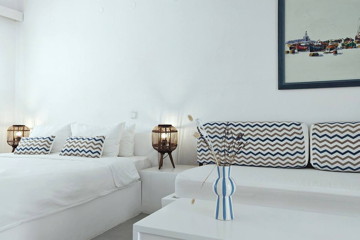 A hotel room with two beds, a white sofa, a small table, and a wall-mounted painting of a harbor scene.