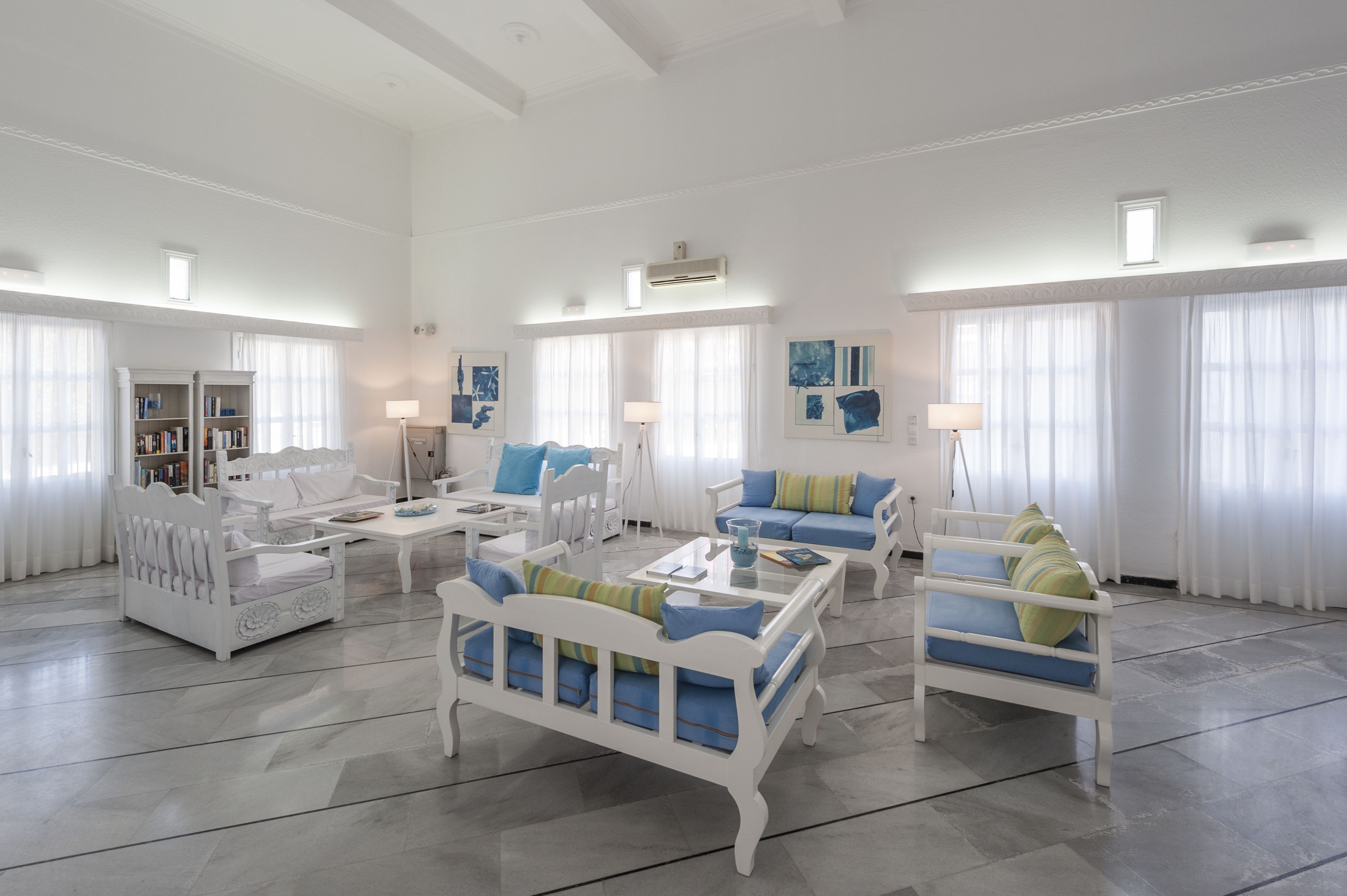 A spacious living room with white furniture, a bookshelf, and large windows with sheer curtains.