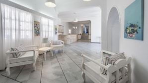 A spacious living area with white furniture, a kitchen in the background, and a large painting on the wall.
