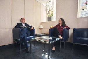 Dos personas sentadas en un área de espera moderna con una mesa de vidrio, una lámpara y un jarrón con flores.