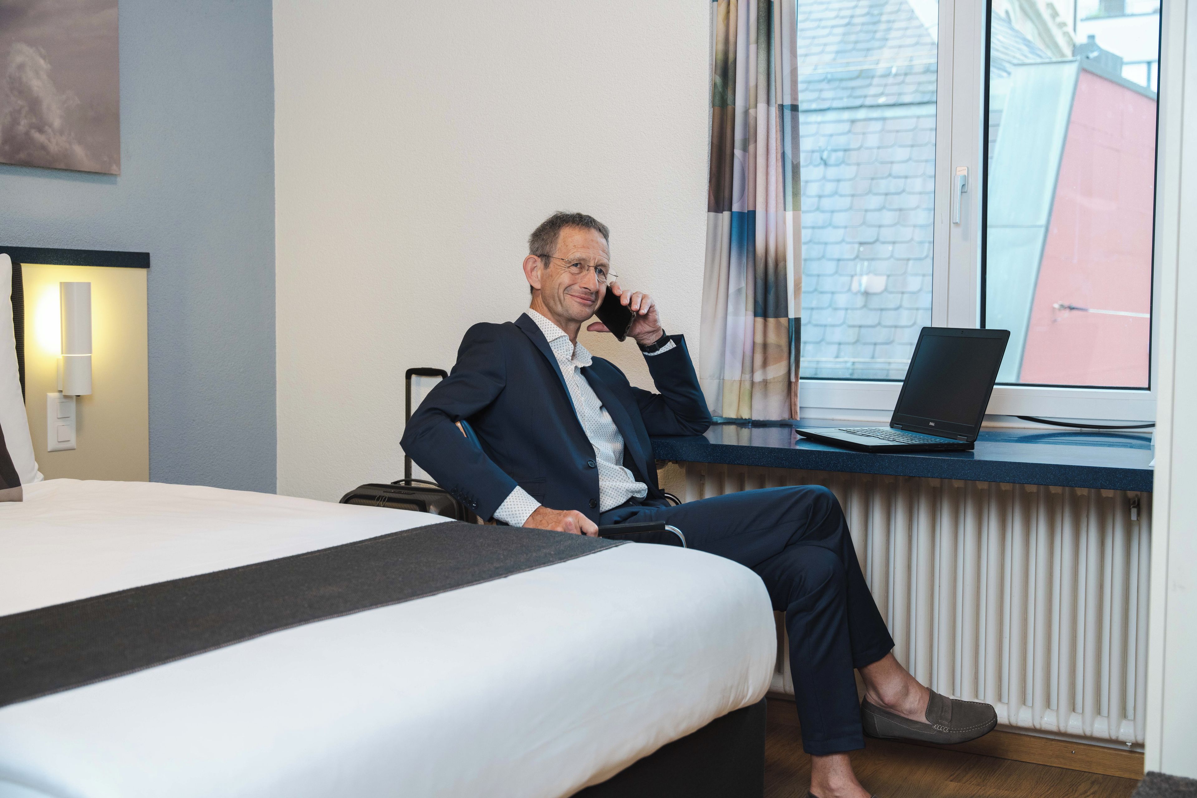 A man in a suit sitting at a desk with a laptop, a bed with a black headboard, and a window with blinds.