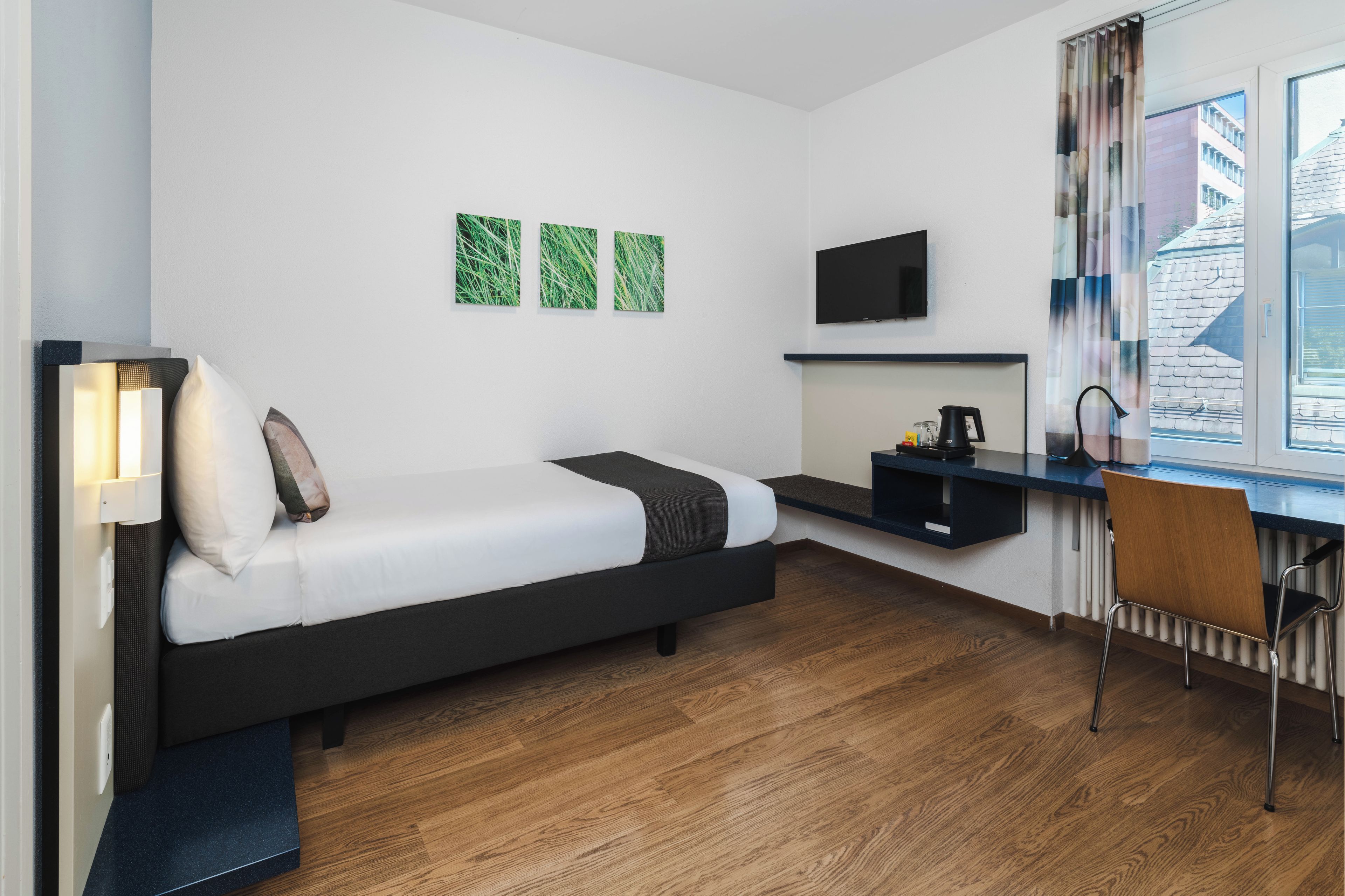 A modern hotel room with a bed, a desk, a chair, a TV, and a window with a view of buildings.
