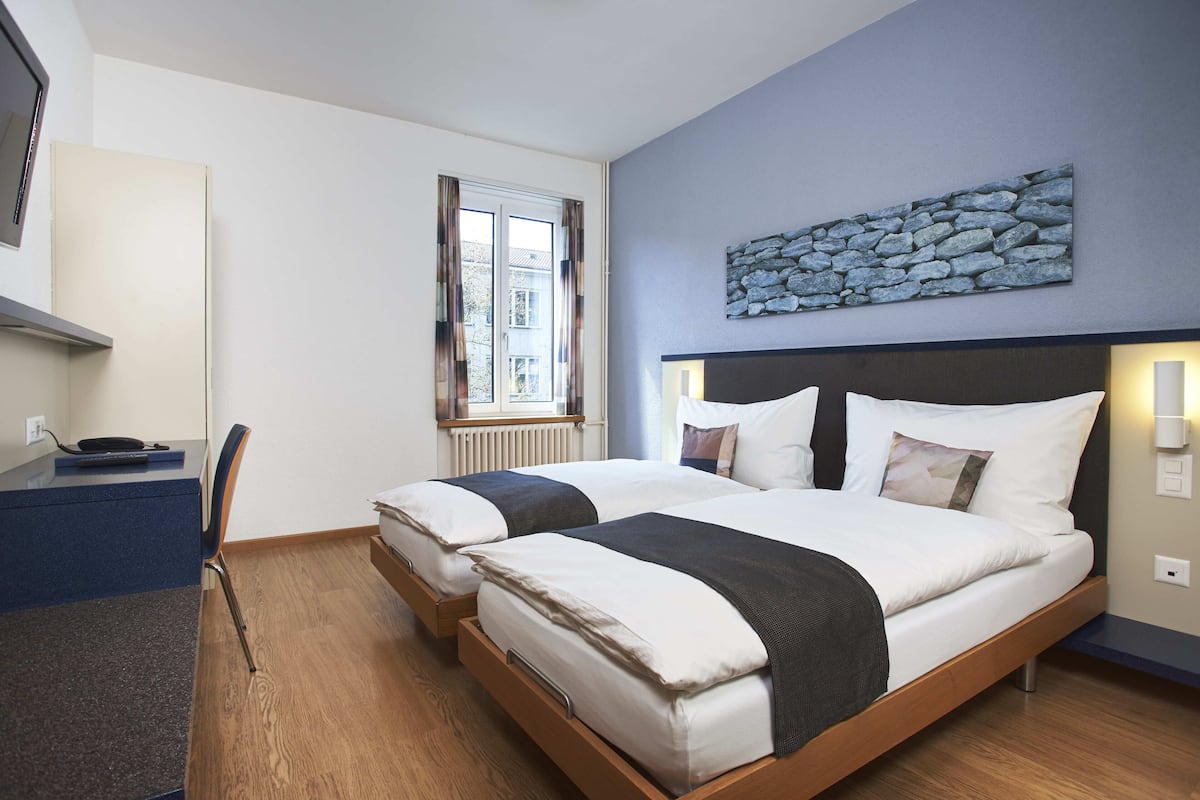 Une chambre d’hôtel avec deux lits, un bureau, une chaise, une fenêtre avec des rideaux et une œuvre d’art murale en pierre.