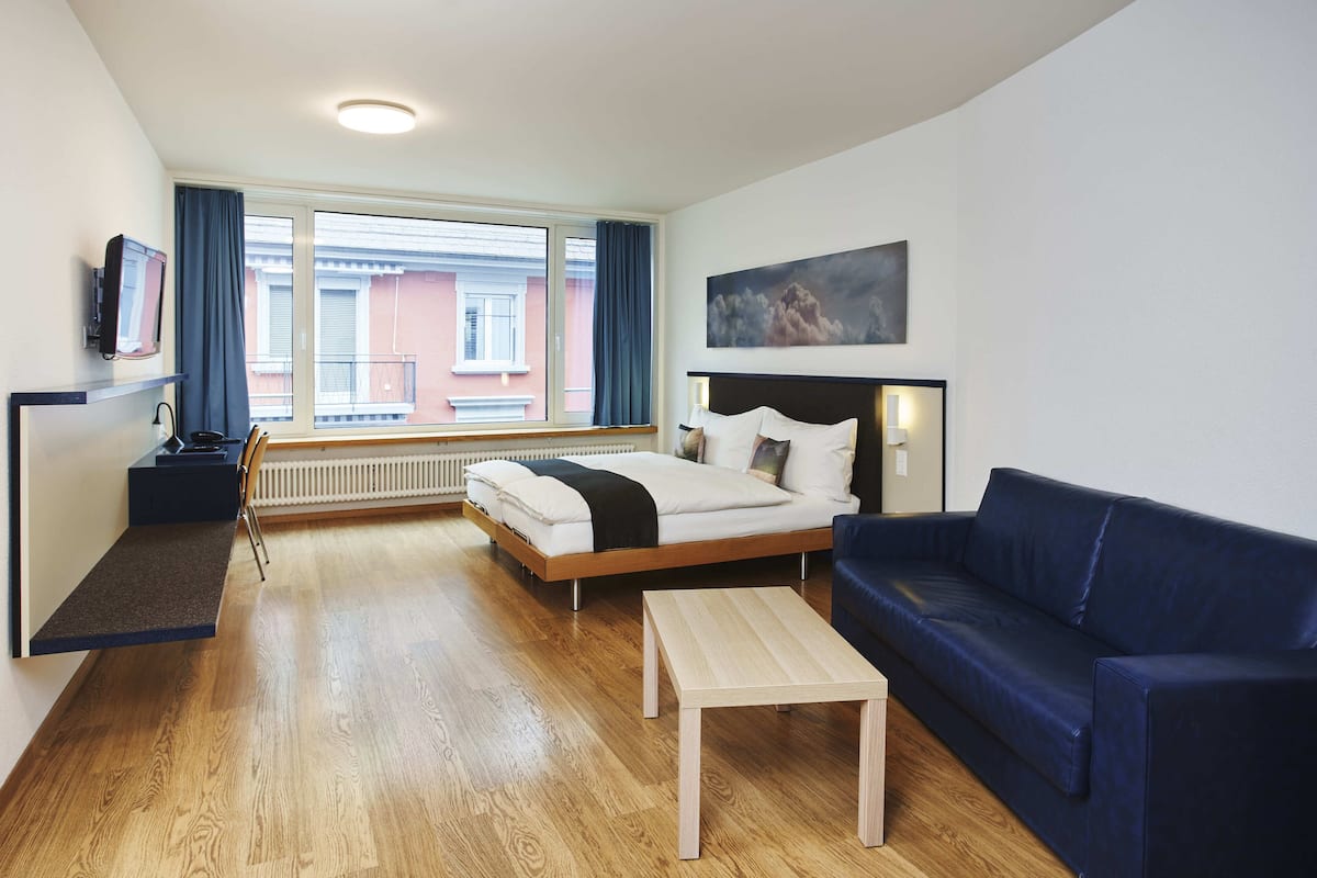 Une chambre d’hôtel avec un lit, un canapé bleu, une table basse en bois, un téléviseur posé sur une étagère et une fenêtre avec des rideaux.
