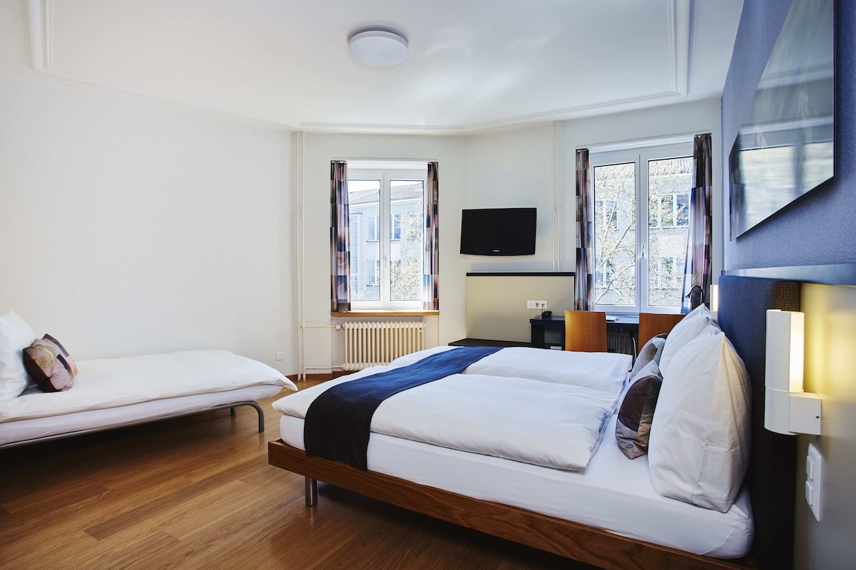 A bedroom with a bed, a smaller bed, a TV, and a window with curtains.