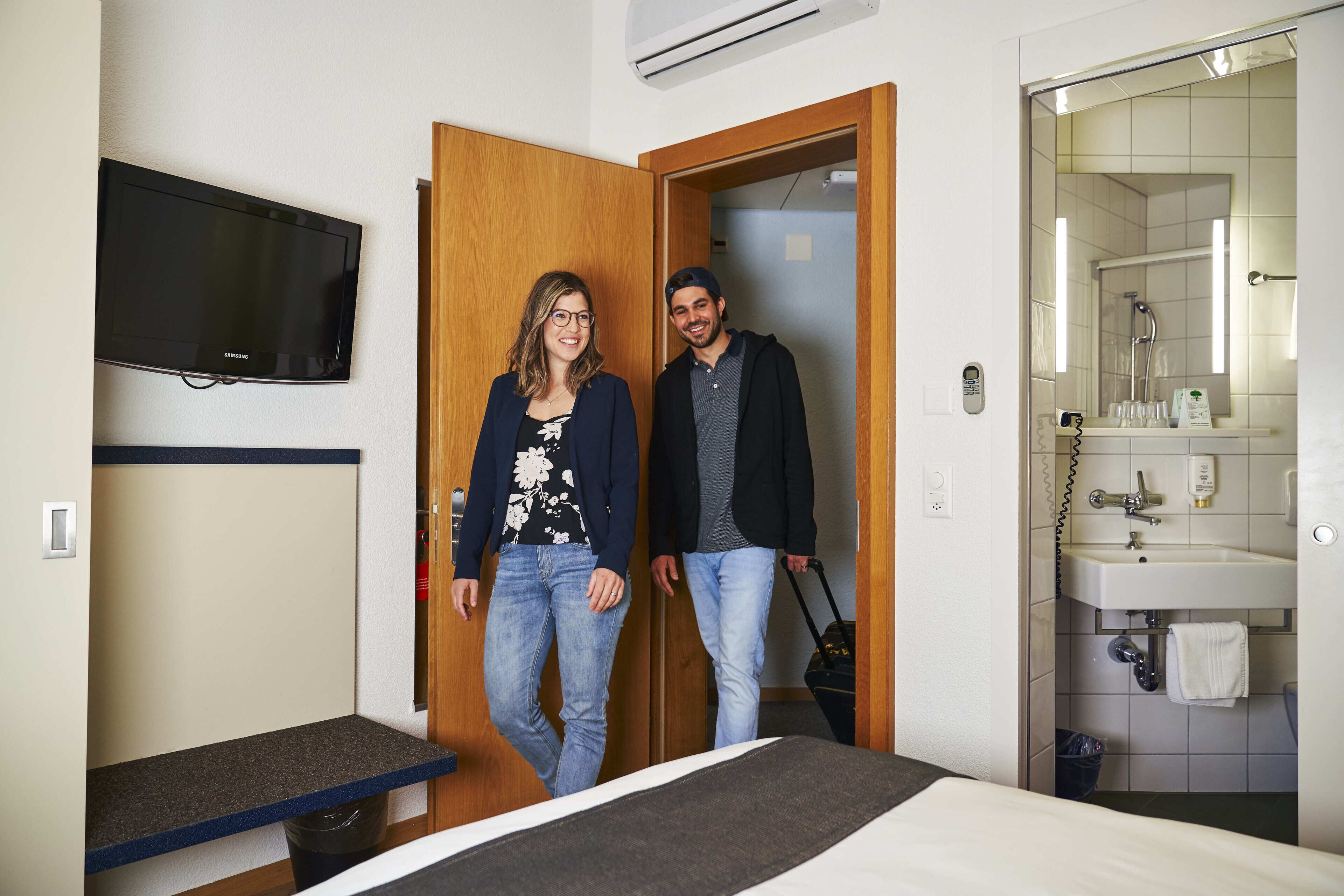 A hotel room with a bed, a desk, a TV mounted on the wall, and a bathroom visible through an open door.