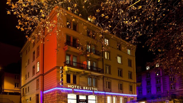 A multi-story hotel with illuminated signage, a visible entrance, and a staircase leading to the entrance. - Bristol Hotel (Zürich)