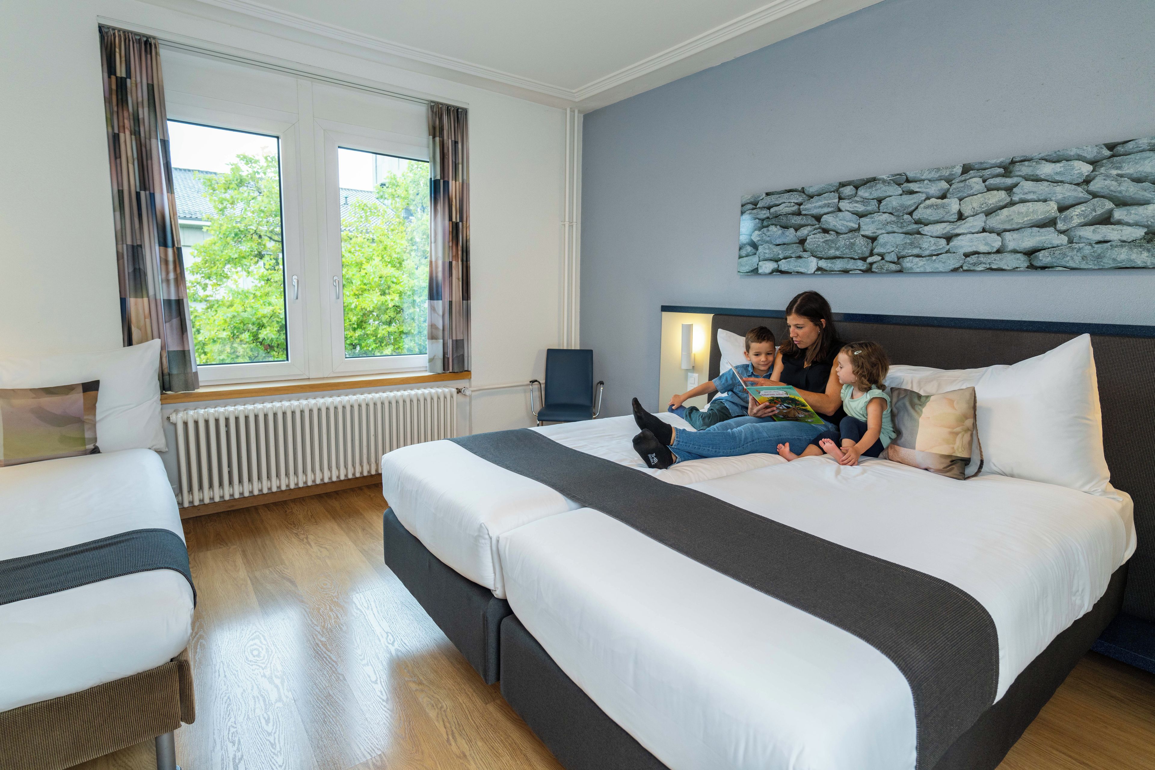 A hotel room with a large bed, a window with curtains, a stone wall decoration, and a radiator.