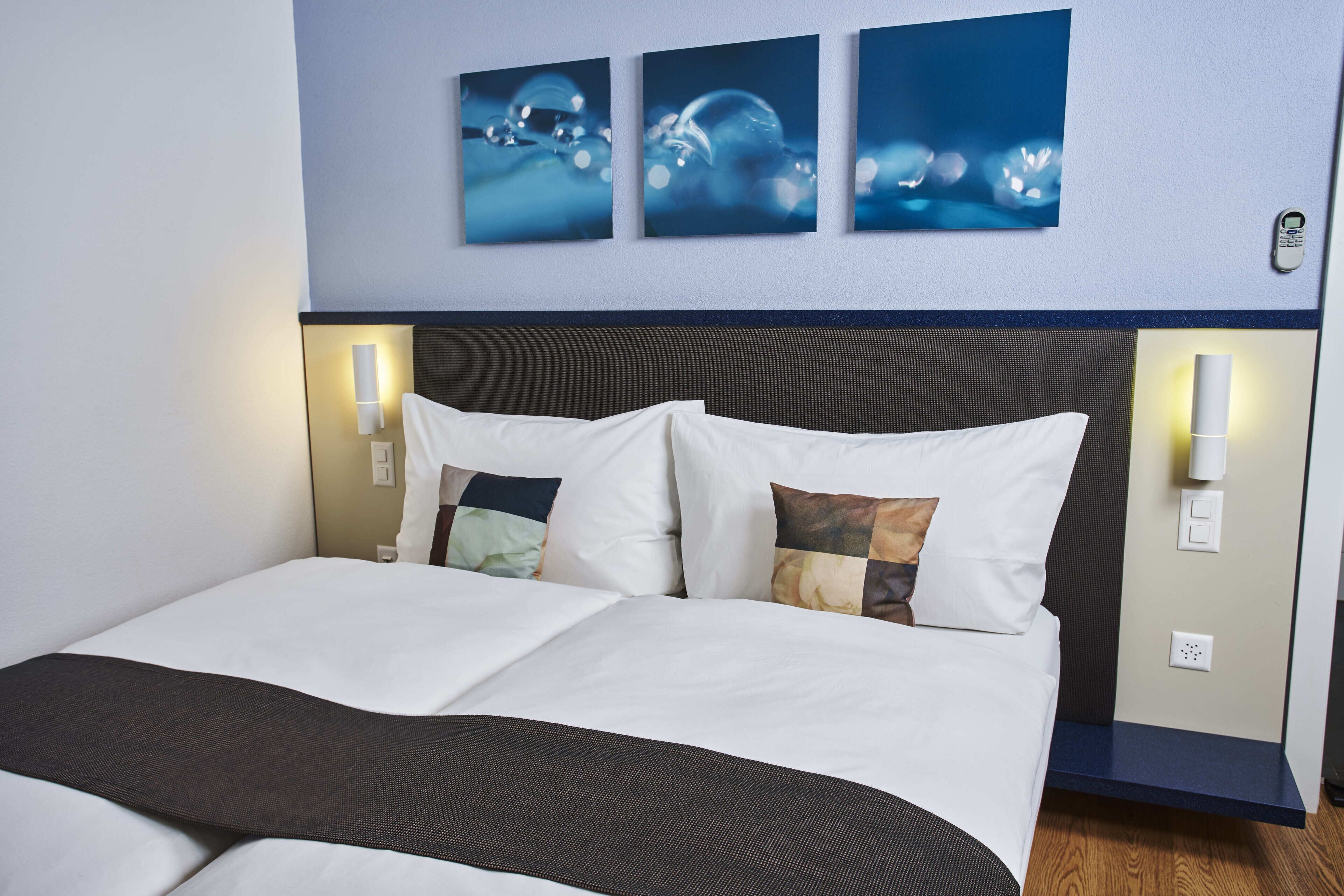 Una cama bien hecha con sábanas blancas y una manta marrón oscuro, con dos lámparas de noche a los lados y un teléfono instalado en la pared.