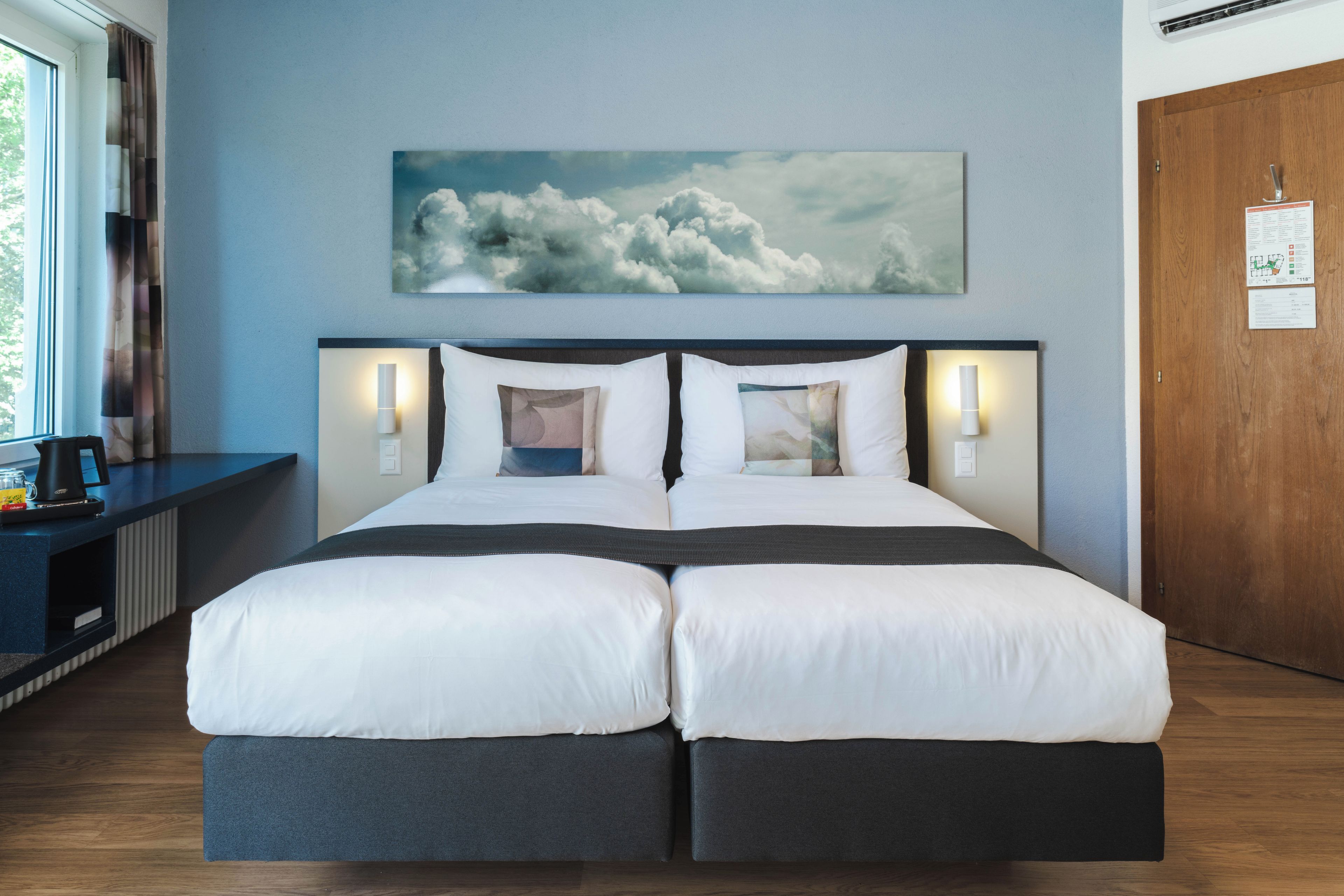 A double bed with a dark headboard and white bedding, a wall-mounted artwork of clouds, and a wooden door with a sign.